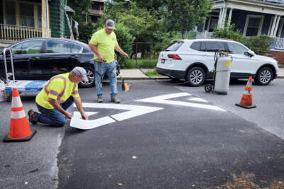 A crew works to adhere markings onto a speed hump on Peter Parley Road in Jamaica Plain. One man places chevron-shaped decals on the hump while another adheres the decal using a blowtorch. (Andrea Perdomo-Hernandez/WBUR)